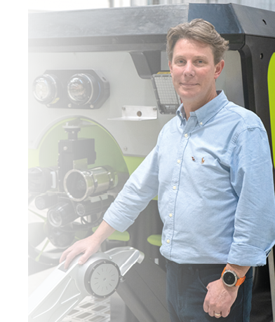 A man wearing a blue shirt and blue jeans is standing in front of a green and white machine. Description generated by AI