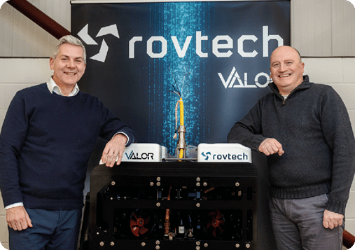 Two men standing in front of a black and white sign. Description generated by AI